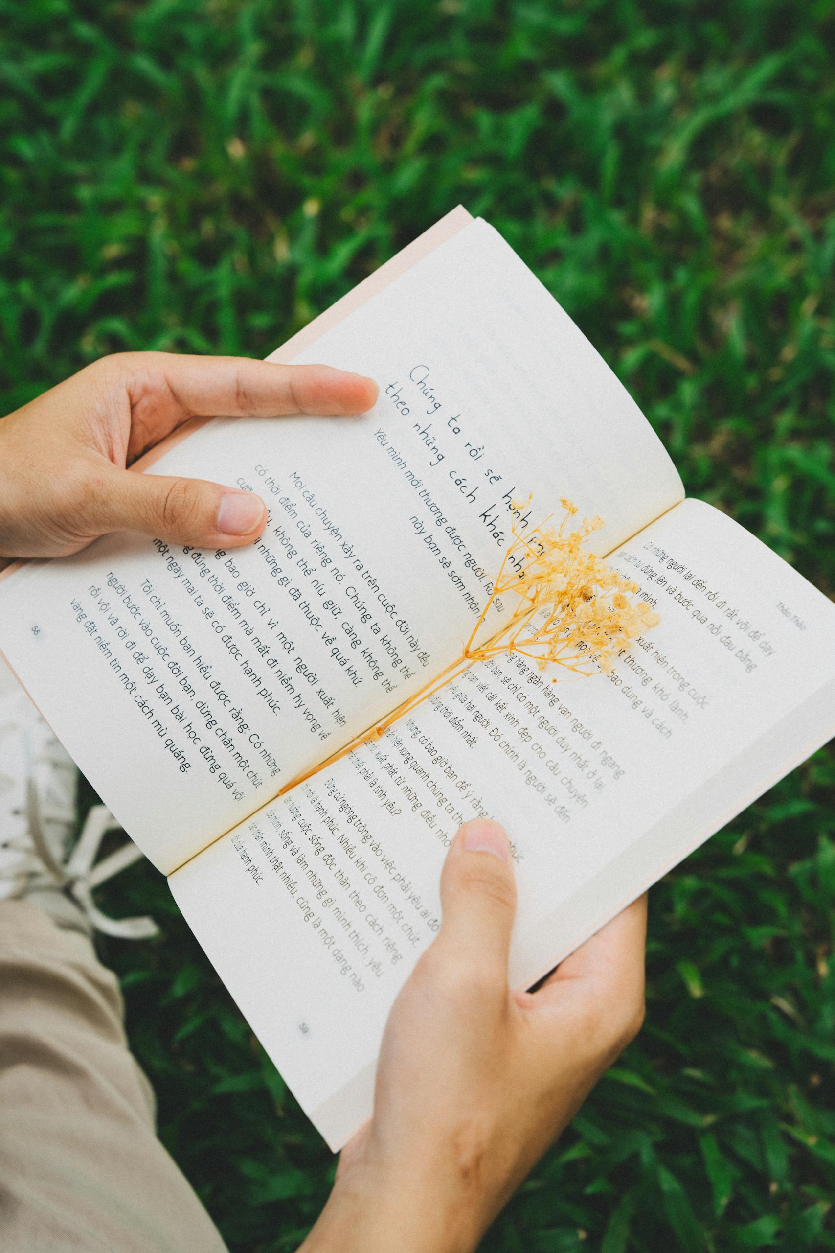 A person is holding an open book with dried flowers, resting on green grass outdoors.